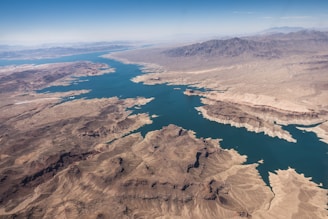 Aerial view of a large, winding body of water cutting through a rugged desert landscape. The terrain features rocky, layered mountains and ridges, with a mix of brown and beige colors. The blue water contrasts sharply with the surrounding arid environment. The horizon is visible in the distance, with clear skies above.