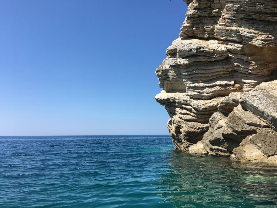 The image depicts a rocky cliff face with a clear blue sky above it, creating a striking contrast between the rugged rock formations and the serene ocean below. The perspective is from the water, looking up at the cliff, which adds depth to the scene. In the distance, there are several birds flying freely in the sky, adding a sense of movement and life to the otherwise still landscape.
