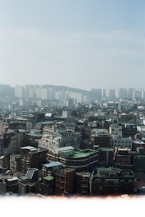 An urban landscape featuring numerous mid-rise buildings with a variety of architectural styles, including flat roofs and rooftop structures. In the background, a line of taller apartment buildings stands against a slightly hazy blue sky. The area appears densely populated, with a mix of residential and commercial buildings.