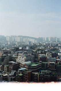 An urban landscape featuring numerous mid-rise buildings with a variety of architectural styles, including flat roofs and rooftop structures. In the background, a line of taller apartment buildings stands against a slightly hazy blue sky. The area appears densely populated, with a mix of residential and commercial buildings.