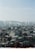 An urban landscape featuring numerous mid-rise buildings with a variety of architectural styles, including flat roofs and rooftop structures. In the background, a line of taller apartment buildings stands against a slightly hazy blue sky. The area appears densely populated, with a mix of residential and commercial buildings.