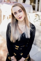 A woman with long, straight blonde hair and bright blue eyes is wearing a black top and a floral skirt. Her makeup is carefully applied with deep red lipstick, and she's accessorized with a necklace and earrings. She stands on a patterned tile floor with a blurred background of outdoor furniture and decor.