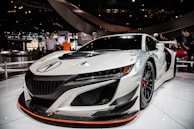 A sleek and modern white sports car with black accents is displayed indoors at an auto show. The car features an aerodynamic design, futuristic headlight aesthetics, and stylish wheels. Several people are observing the car in the background, and the setting is well-lit with shiny surfaces reflecting lights.