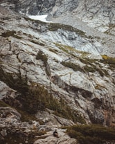 A rugged mountain landscape with large rocky surfaces and patches of green vegetation. The scene is dramatic, with steep cliffs and a distant patch of snow near the top. A person is sitting on the rocks in the lower part of the image, suggesting a sense of exploration and adventure.