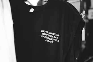 A black t-shirt with white text that reads 'YOU'RE MORE THAN WHAT THEY SAY, YOU'RE BEAUTIFUL & BRAVE' is displayed. The shirt appears to be hanging in a dimly lit area, with parts of another garment visible beside it.