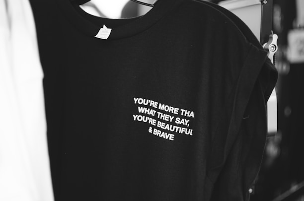A black t-shirt with white text that reads 'YOU'RE MORE THAN WHAT THEY SAY, YOU'RE BEAUTIFUL & BRAVE' is displayed. The shirt appears to be hanging in a dimly lit area, with parts of another garment visible beside it.