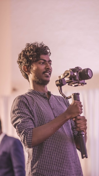 A person holding and operating a professional camera mounted on a stabilizer, focused on capturing footage. The person is wearing a checkered shirt and has curly hair. The background is softly blurred, highlighting the subject.