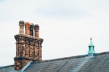 A brick chimney with four terracotta chimney pots is situated on the roof of a building, alongside a small metal turret with a pointed roof. The sky is overcast, providing a soft light over the scene, with the grey slate tiles of the roof complementing the brickwork and metal details.