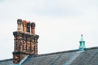 A brick chimney with four terracotta chimney pots is situated on the roof of a building, alongside a small metal turret with a pointed roof. The sky is overcast, providing a soft light over the scene, with the grey slate tiles of the roof complementing the brickwork and metal details.