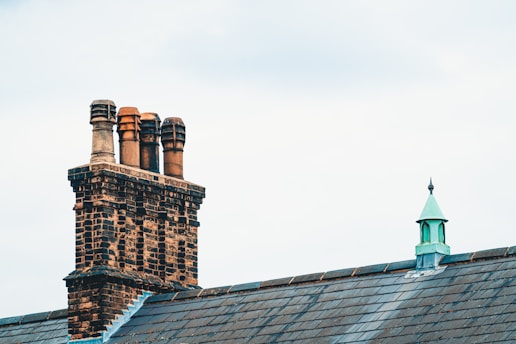 A brick chimney with four terracotta chimney pots is situated on the roof of a building, alongside a small metal turret with a pointed roof. The sky is overcast, providing a soft light over the scene, with the grey slate tiles of the roof complementing the brickwork and metal details.