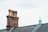 A brick chimney with four terracotta chimney pots is situated on the roof of a building, alongside a small metal turret with a pointed roof. The sky is overcast, providing a soft light over the scene, with the grey slate tiles of the roof complementing the brickwork and metal details.