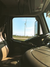 The interior of a vehicle, likely a truck, captures a view through the window showcasing a scenic field with several wind turbines. The front seat and steering wheel are visible, suggesting the perspective from the driver's side. The sky is clear with a few clouds, adding to the serene landscape.