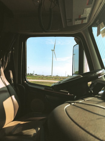 The interior of a vehicle, likely a truck, captures a view through the window showcasing a scenic field with several wind turbines. The front seat and steering wheel are visible, suggesting the perspective from the driver's side. The sky is clear with a few clouds, adding to the serene landscape.