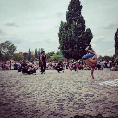 A street performer is engaged in a breakdancing move, balancing on one hand in an outdoor setting. A crowd of spectators surrounds him, some sitting and others standing, watching the performance. The scene is set on a cobblestone courtyard with tall, leafy trees in the background under a cloudy sky.