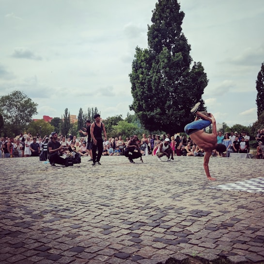 A street performer is engaged in a breakdancing move, balancing on one hand in an outdoor setting. A crowd of spectators surrounds him, some sitting and others standing, watching the performance. The scene is set on a cobblestone courtyard with tall, leafy trees in the background under a cloudy sky.