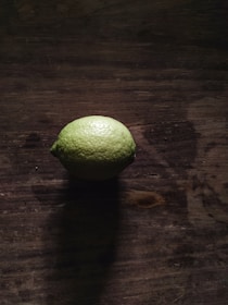 A single lime sits on a dark, rustic wooden surface, casting a shadow. The lighting is moody, with highlights emphasizing the texture of both the lime and the wood.