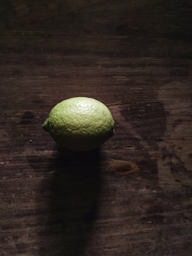 A single lime sits on a dark, rustic wooden surface, casting a shadow. The lighting is moody, with highlights emphasizing the texture of both the lime and the wood.