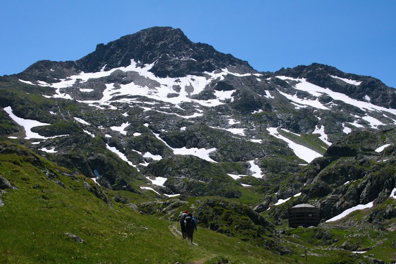 A rugged mountain landscape with patches of snow scattered across the rocky terrain. In the foreground, a hiker is making their way along a grassy trail, surrounded by lush green slopes. The sky is clear and blue, adding to the sense of openness and tranquility.