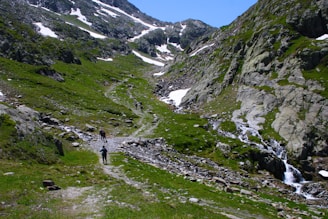 A mountainous landscape features a narrow path leading through lush green grass and patches of snow. Several hikers are seen walking along the trail, surrounded by rocky terrain and a clear blue sky. A small waterfall cascades down the rocks on the right side.