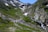 A mountainous landscape features a narrow path leading through lush green grass and patches of snow. Several hikers are seen walking along the trail, surrounded by rocky terrain and a clear blue sky. A small waterfall cascades down the rocks on the right side.