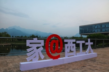 A large outdoor sign features a combination of large white characters and a prominent red at symbol (@), positioned on a wooden platform. In the background, there is a tranquil body of water with lush greenery and distant mountains. A modern glass building is visible to the right.