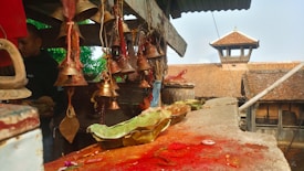 An assortment of brass bells hangs from a wooden structure, creating a spiritual atmosphere. The surface below is stained with red powder and adorned with green leaves, likely used in a ritual. A rustic building with a tiled roof is visible in the background. A person stands to the left, partially obscured.