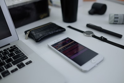 A neatly arranged collection of everyday items on a white surface, including a laptop, smartphone displaying a colorful app interface, a black woven wallet, a watch with a black strap, a black pen, and a black cup.
