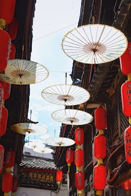 Traditional Chinese paper umbrellas hang amidst a narrow alley lined with vibrant red lanterns. Sunlight filters through the umbrellas, casting delicate shadows. Architectural details of classic Chinese wooden structures are visible on either side, adding to the cultural ambiance.