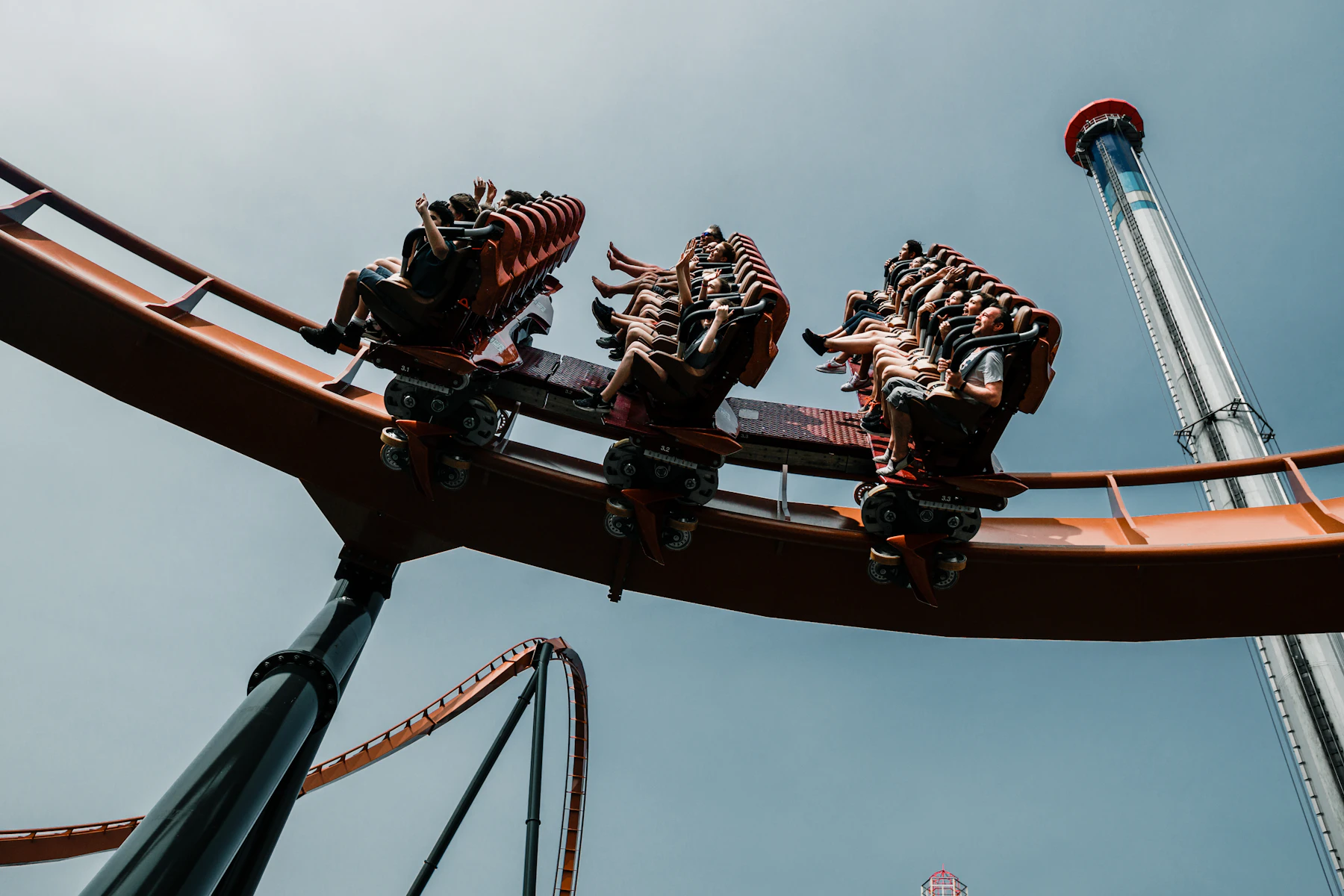 Seen from below, riders in over-the-shoulder restraints on an orange steel coaster train with their legs hanging free, a drop tower rising behind