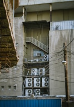 A building's exterior features multiple air conditioning units stacked and mounted on metal racks near a fire escape. The facade is weathered, with visible electrical wires crisscrossing the scene. An adjacent structure with a wooden overhang is partially visible.