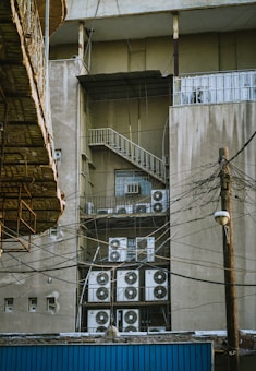 A building's exterior features multiple air conditioning units stacked and mounted on metal racks near a fire escape. The facade is weathered, with visible electrical wires crisscrossing the scene. An adjacent structure with a wooden overhang is partially visible.