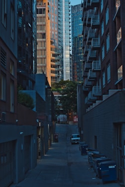 A narrow urban alley flanked by tall buildings with glass and brick facades. The alley is lined with dumpsters on one side, and a single parked car is visible. The buildings rise on either side, showcasing modern architecture with numerous balconies and large windows. In the distance, trees and a sign with the word 'LOCAL' can be seen.