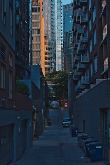 A narrow urban alley flanked by tall buildings with glass and brick facades. The alley is lined with dumpsters on one side, and a single parked car is visible. The buildings rise on either side, showcasing modern architecture with numerous balconies and large windows. In the distance, trees and a sign with the word 'LOCAL' can be seen.