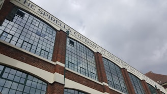 A large brick building with numerous rectangular windows is seen under an overcast sky. The building features a sign at the top reading 'The Spirella Company of Great Britain LTD - High Grade Corsets'. The architecture suggests an industrial or commercial purpose with a historic aspect.