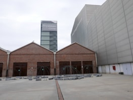 A contemporary architectural scene features a modern glass tower labeled with 'Prysmian Group' in the background, adjacent to an industrial-style building with a brick façade and multiple girded entrances. The area in front includes geometric art installations or sculptures surrounded by concrete blocks on a wide paved surface.