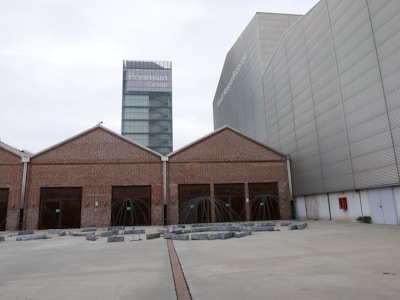 A contemporary architectural scene features a modern glass tower labeled with 'Prysmian Group' in the background, adjacent to an industrial-style building with a brick façade and multiple girded entrances. The area in front includes geometric art installations or sculptures surrounded by concrete blocks on a wide paved surface.
