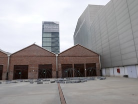 A contemporary architectural scene features a modern glass tower labeled with 'Prysmian Group' in the background, adjacent to an industrial-style building with a brick façade and multiple girded entrances. The area in front includes geometric art installations or sculptures surrounded by concrete blocks on a wide paved surface.