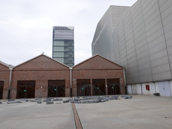 A contemporary architectural scene features a modern glass tower labeled with 'Prysmian Group' in the background, adjacent to an industrial-style building with a brick façade and multiple girded entrances. The area in front includes geometric art installations or sculptures surrounded by concrete blocks on a wide paved surface.