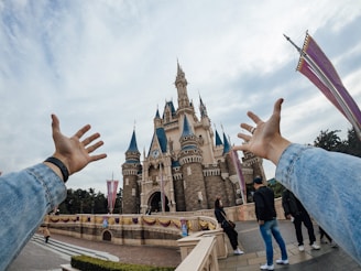 A majestic castle with tall spires under a partly cloudy sky, viewed from a first-person perspective with arms outstretched towards it. People are casually strolling in the foreground, dressed in casual clothing. The scene conveys a sense of excitement and wonder.