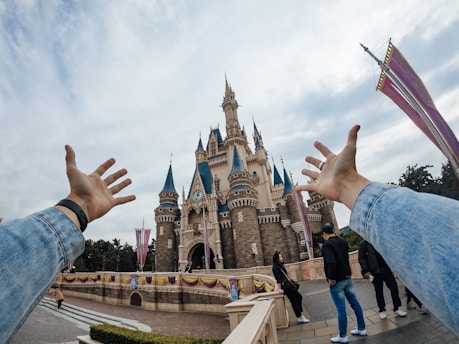 A majestic castle with tall spires under a partly cloudy sky, viewed from a first-person perspective with arms outstretched towards it. People are casually strolling in the foreground, dressed in casual clothing. The scene conveys a sense of excitement and wonder.