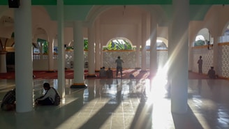 Sunlight streams through the arches of a mosque’s interior, casting long shadows on the polished tiled floor. Several people are seated or standing on the red carpeted area, likely engaged in personal reflection or prayer. The architecture features elegant columns and latticework.