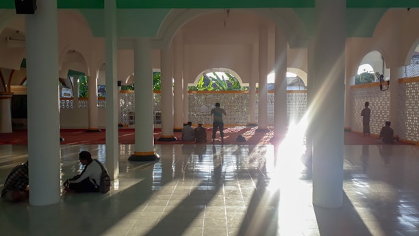 Sunlight streams through the arches of a mosque’s interior, casting long shadows on the polished tiled floor. Several people are seated or standing on the red carpeted area, likely engaged in personal reflection or prayer. The architecture features elegant columns and latticework.