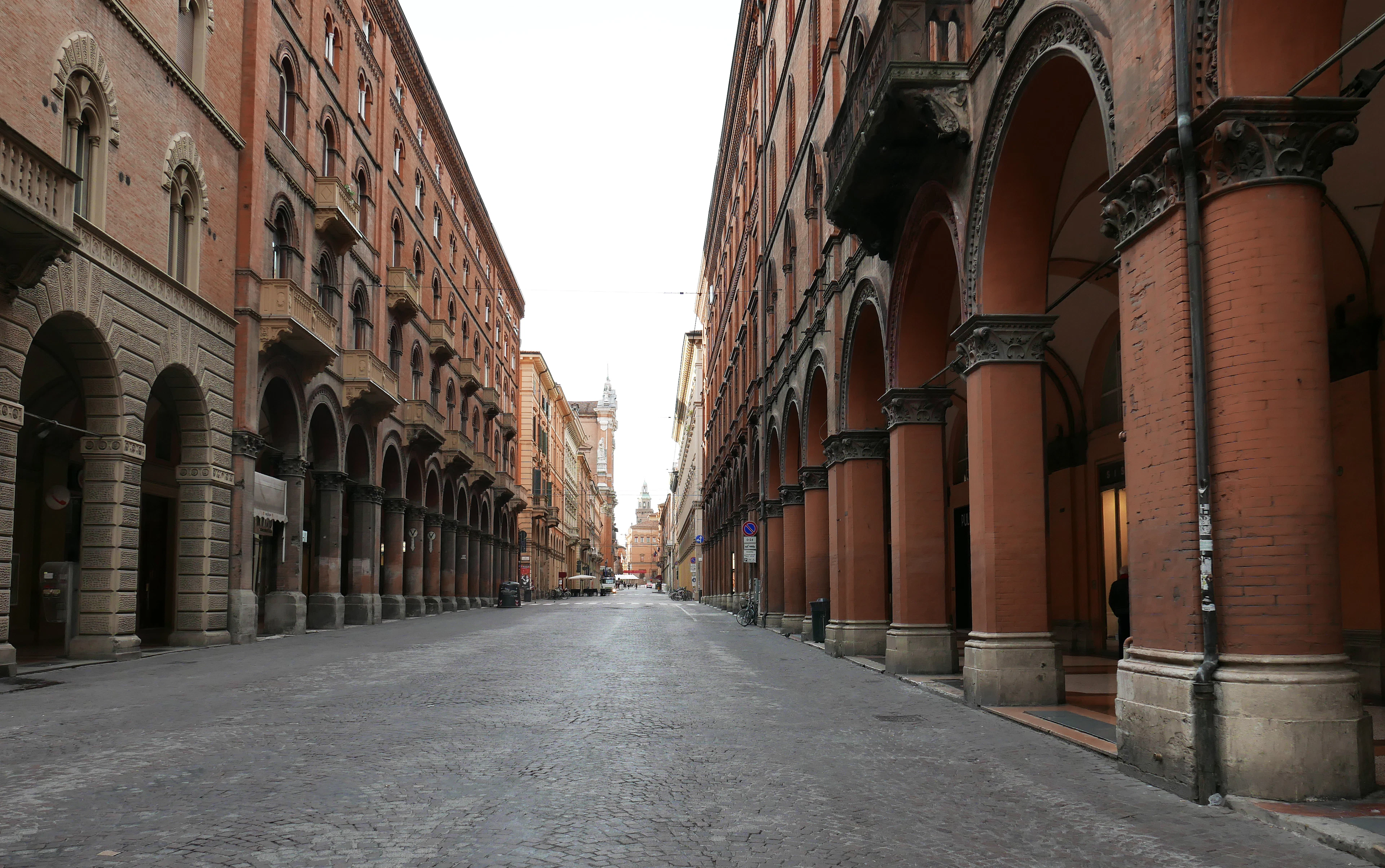 An empty red-brick portico corridor in Bologna stretching toward a distant medieval tower.