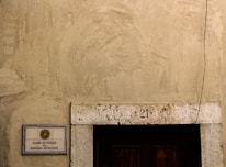 A weathered exterior wall with a rough texture, featuring a rectangular sign on the left side displaying 'CLUBE DE OFICIAIS DA MARINHA MERCANTE'. Above a dark wooden door is the number '21' inscribed on a stone lintel, and a single cable runs vertically down the right side.