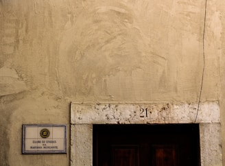 A weathered exterior wall with a rough texture, featuring a rectangular sign on the left side displaying 'CLUBE DE OFICIAIS DA MARINHA MERCANTE'. Above a dark wooden door is the number '21' inscribed on a stone lintel, and a single cable runs vertically down the right side.