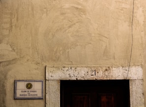 A weathered exterior wall with a rough texture, featuring a rectangular sign on the left side displaying 'CLUBE DE OFICIAIS DA MARINHA MERCANTE'. Above a dark wooden door is the number '21' inscribed on a stone lintel, and a single cable runs vertically down the right side.