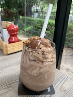 A thick, creamy chocolate milkshake served in a glass with a wide white straw. The top is adorned with chocolate whipped cream and what appears to be a dollop of chocolate syrup or fudge. The glass is placed on a wooden table with a coaster beneath it, near a window with greenery outside. In the background, there is a wooden box holding a red pepper mill and other condiments.