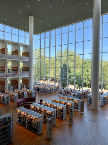 A spacious, modern library interior with tall ceilings and large glass windows allowing natural light to flood the space. Rows of bookshelves are neatly organized on a polished wooden floor. The area features a mix of seating arrangements, including long tables with chairs and curved sofas. Outside the windows, a view of greenery and trees adds a serene backdrop to the setting.