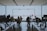 People are sitting and working at tables with laptops inside a store or office space with a large glass window overlooking an outdoor area. A few individuals are standing and interacting near the entrance, while two people are seen outside looking at the view.