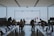 People are sitting and working at tables with laptops inside a store or office space with a large glass window overlooking an outdoor area. A few individuals are standing and interacting near the entrance, while two people are seen outside looking at the view.